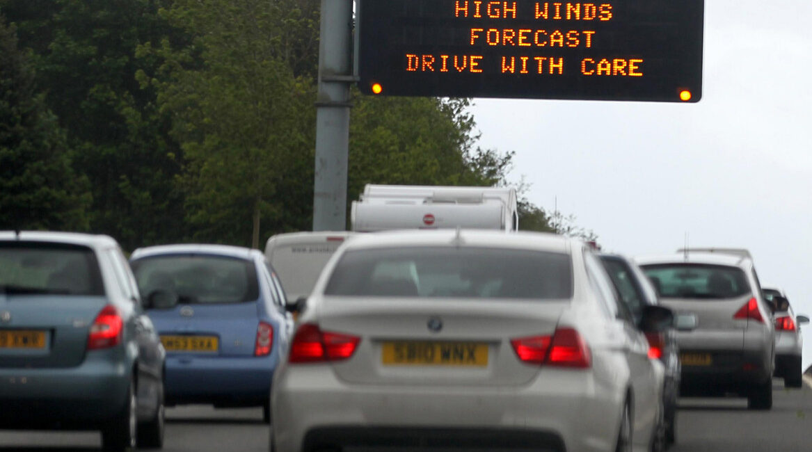 A high-sided Warrington hire van being driven carefully on an exposed bridge during windy weather, with the driver maintaining a firm grip on the steering wheel.