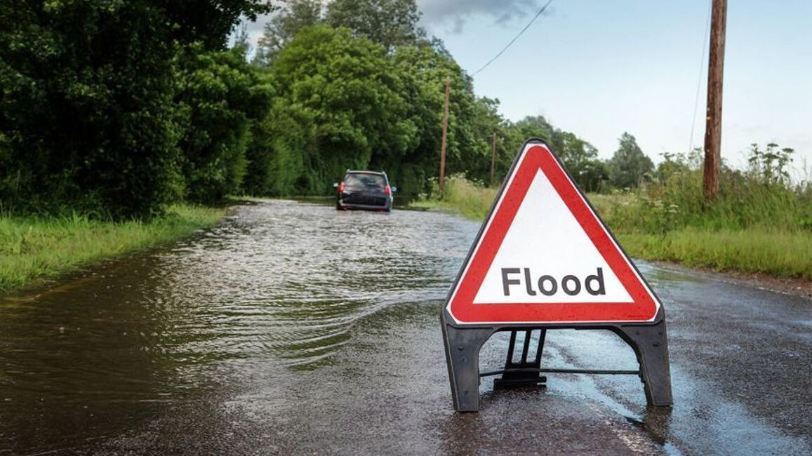 A van driving cautiously through shallow water on flooded roads, creating a small bow wave to protect the engine while maintaining a steady, slow speed.