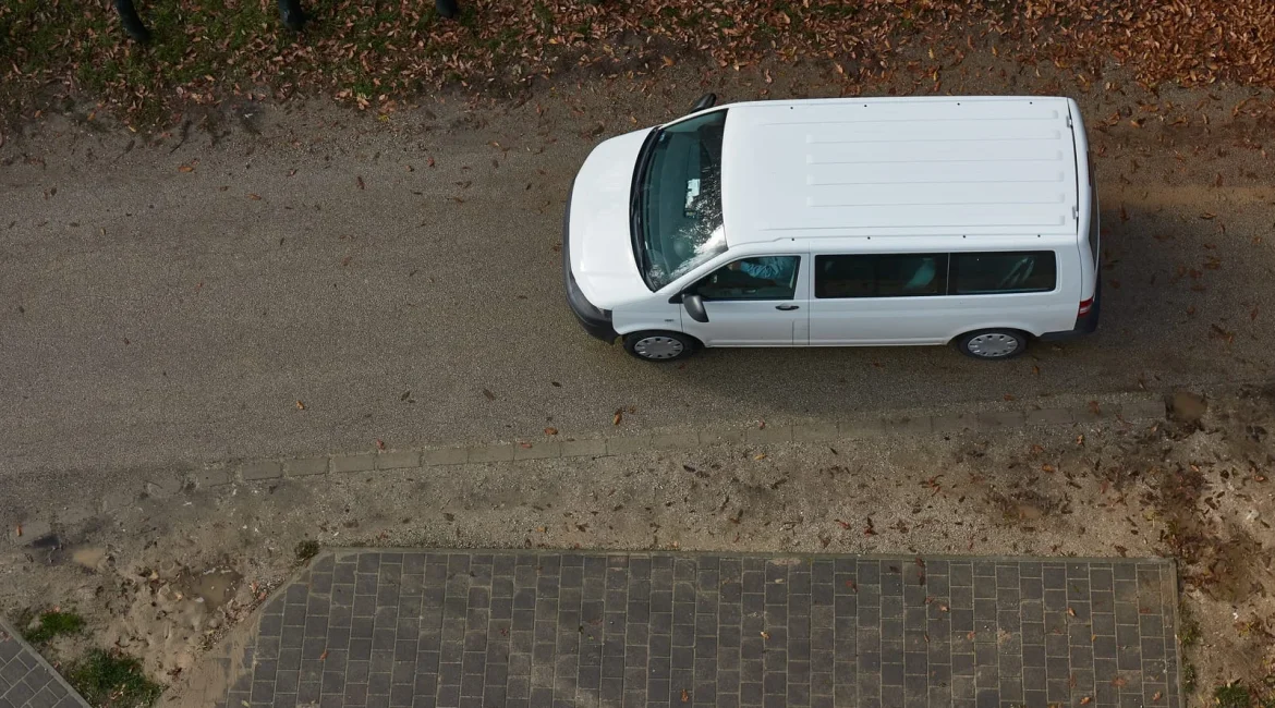 Top-down view of a white van parked on a gravel road beside a brick sidewalk, with fallen autumn leaves nearby.