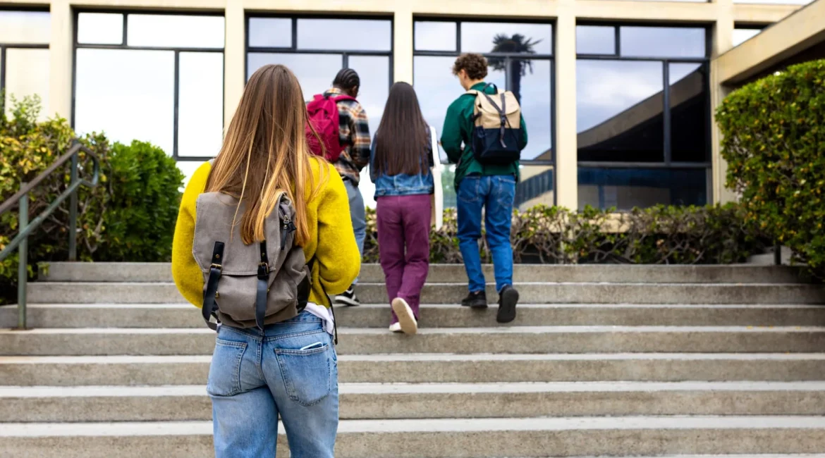Group of students with backpacks walking up concrete stairs toward a modern glass-fronted building, seen from behind.