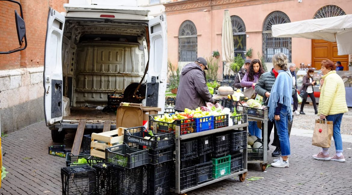 Open white delivery van at a market with crates of fresh vegetables and shoppers examining produce in daylight.